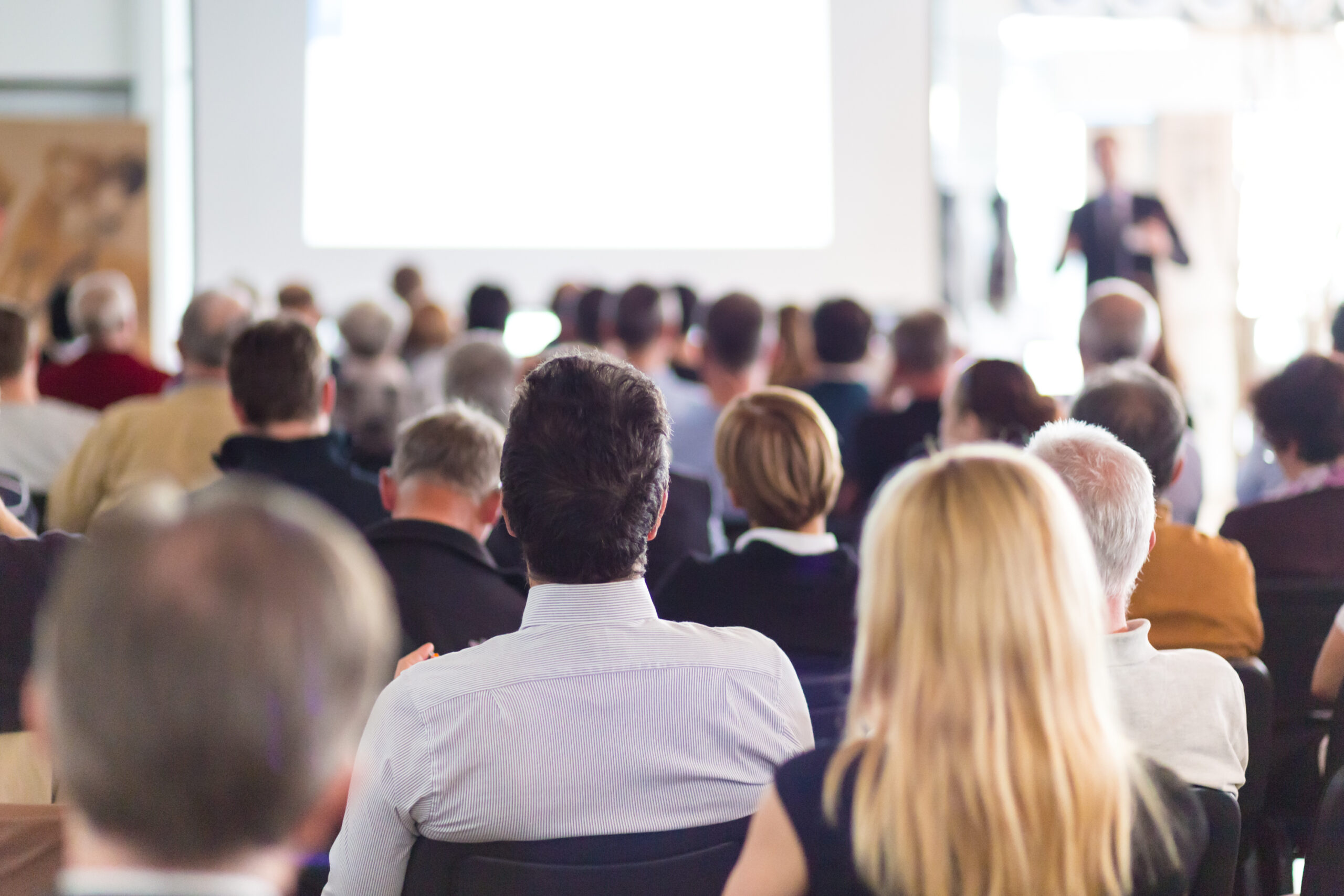 Audience members attentively watching a presentation in a conference setting. The focus is on the speaker in the background.