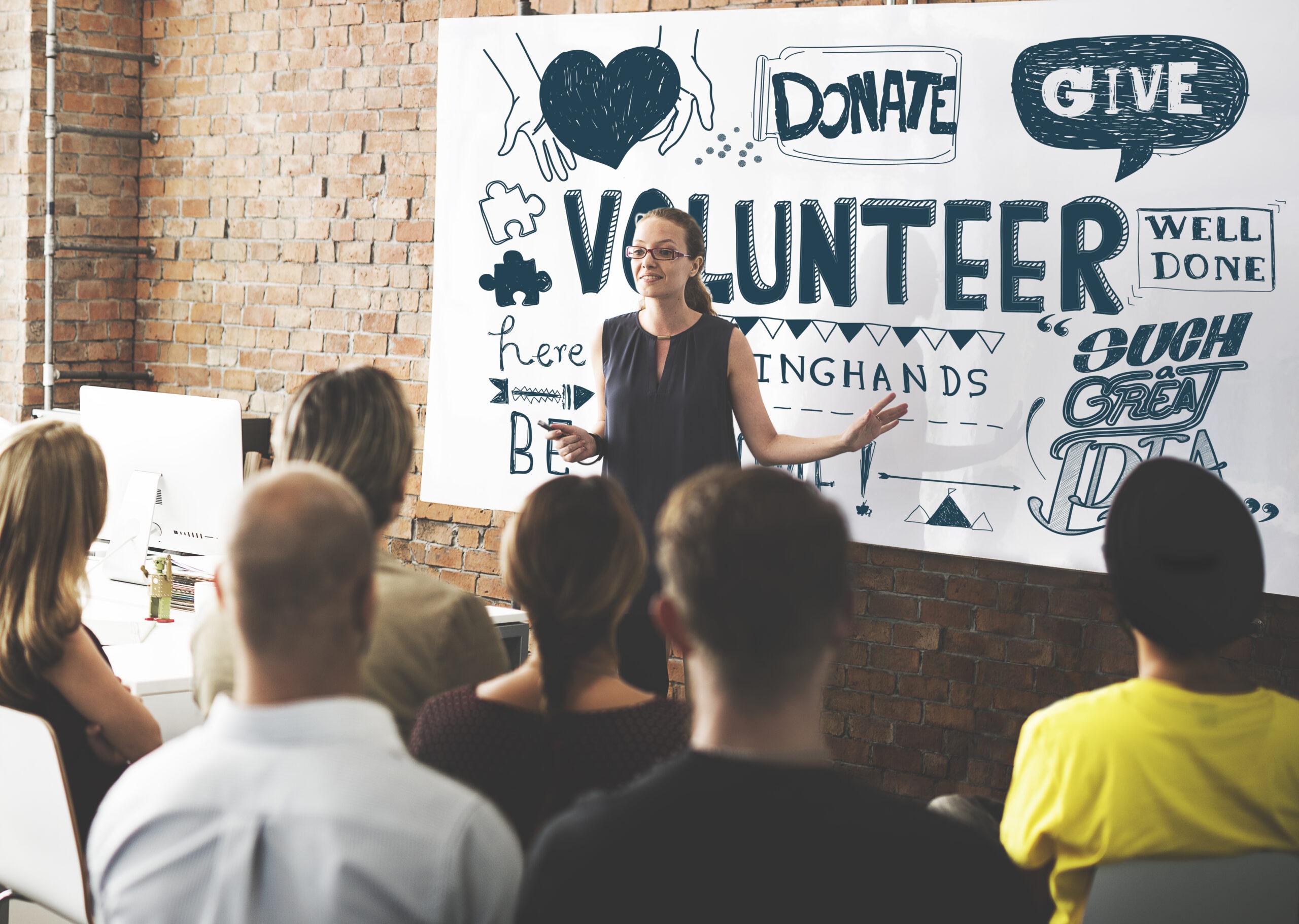 A speaker presents to an engaged audience in a workspace, emphasizing volunteerism with a large backdrop featuring motivational phrases like "Donate" and "Volunteer."