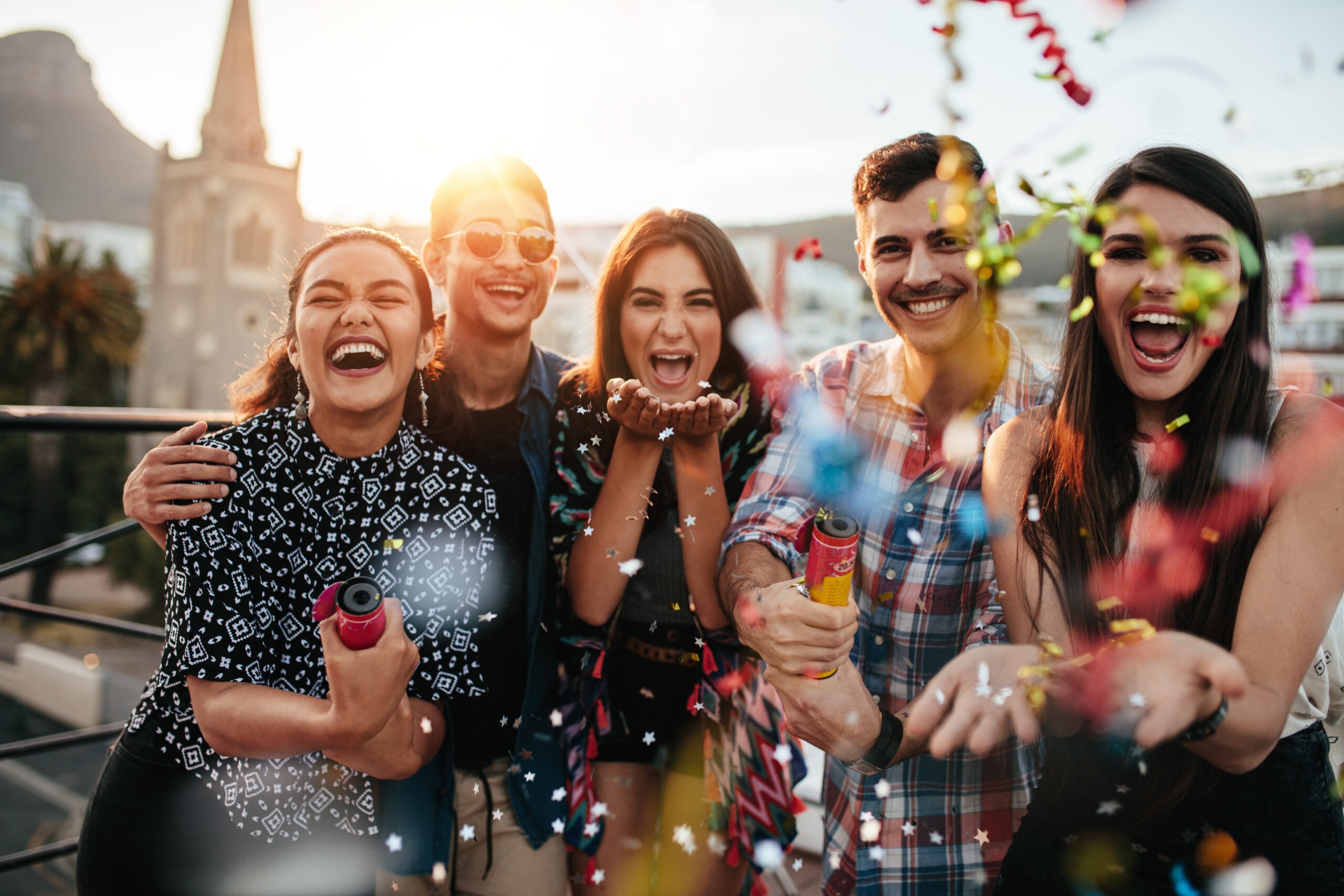 A group of five friends joyfully celebrating outdoors, throwing colorful confetti with smiles and laughter, against a scenic backdrop.