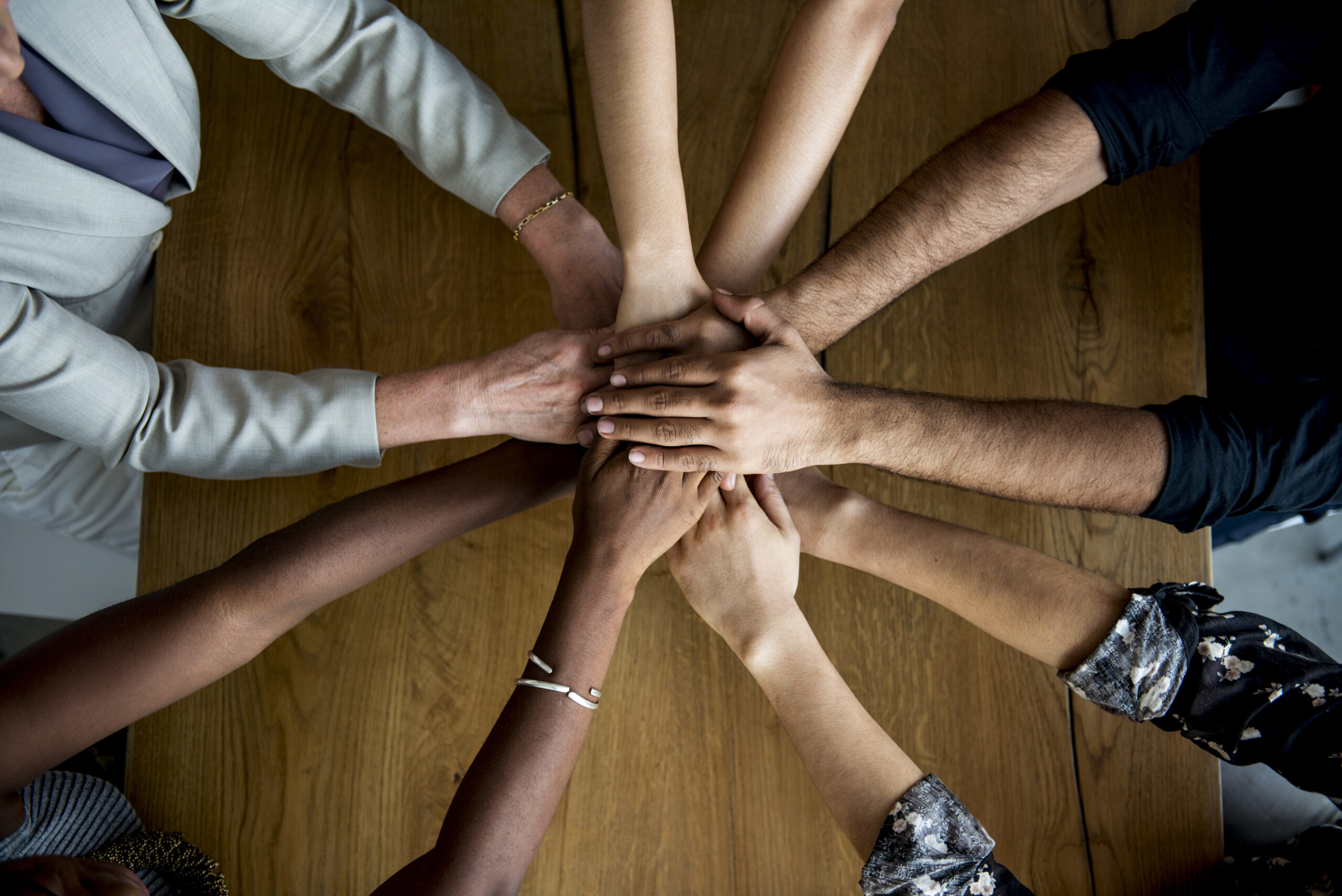 Diverse hands of multiple individuals stack together on a wooden table, symbolizing teamwork and collaboration.
